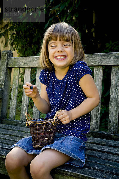 Grinsendes kleines Mädchen sitzt auf einer Gartenbank mit einem Korb voller Blaubeeren.