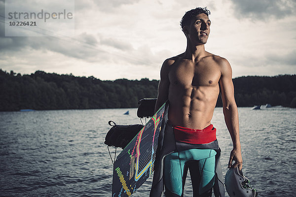Junger Wakeboarder mit seiner Ausrüstung am Seeufer stehend