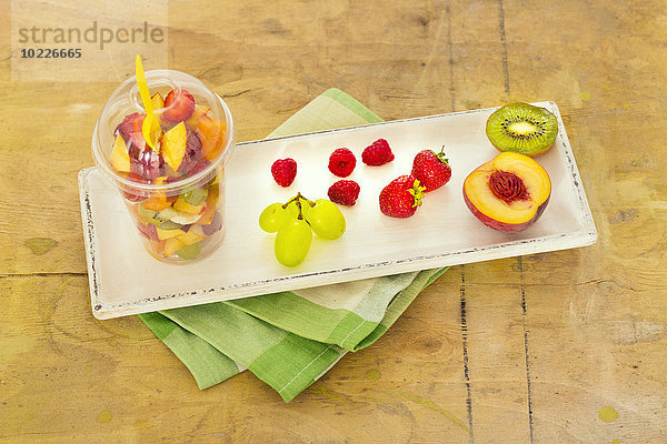 Plastikbecher mit Obstsalat und Früchten auf Holztablett