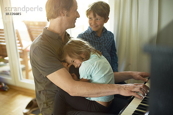 Vater spielt Klavier mit Tochter auf dem Schoß  Sohn schaut zu