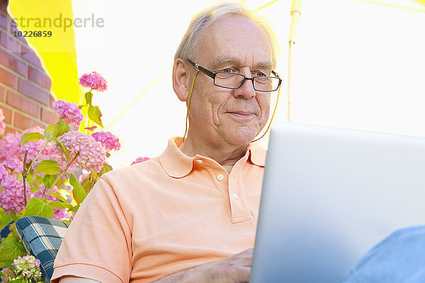 Senior Mann sitzt auf einer Terrasse unter einem Sonnenschirm mit Laptop
