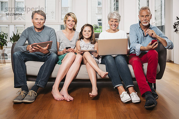 Gruppenbild von drei Generationen Familie mit verschiedenen digitalen Geräten auf einer Couch
