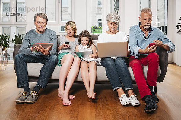 Gruppenbild von drei Generationen Familie  die auf einer Couch mit verschiedenen digitalen Geräten sitzt.