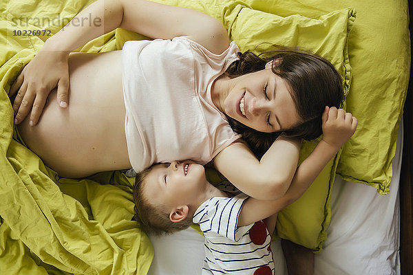 Schwangere Mutter und ihr Sohn kuscheln sich zu Hause im Bett.