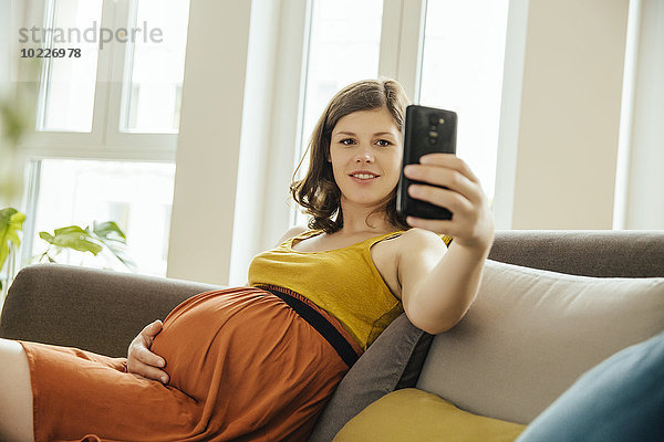 Schwangere Frau  die zu Hause einen Selfie mit Smartphone auf der Couch hat.