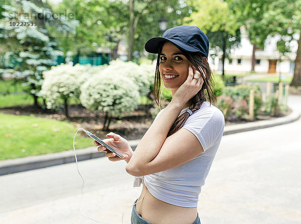 Porträt einer lächelnden jungen Frau beim Musikhören mit Kopfhörern im Park