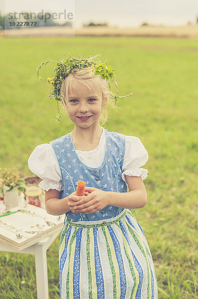 Deutschland  Sachsen  Porträt des lächelnden Mädchens mit Blumenkranz und Dirndl mit Karotte in der Hand