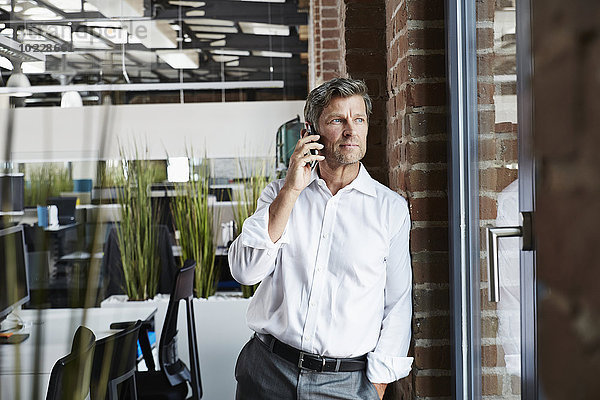 Geschäftsmann im Büro auf dem Smartphone mit Blick aus dem Fenster