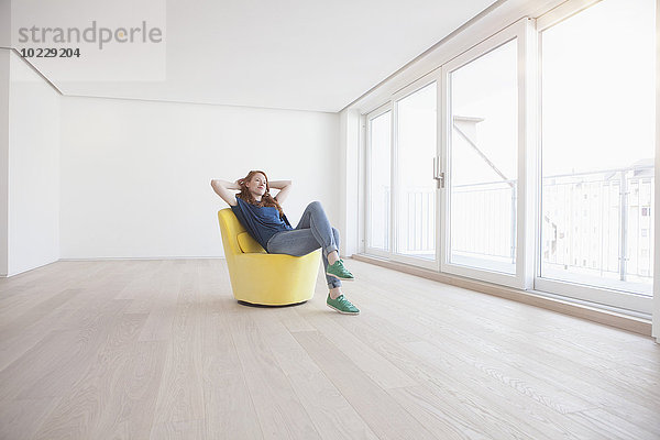 Junge Frau sitzt auf einem gelben Sessel in ihrem leeren Wohnzimmer.