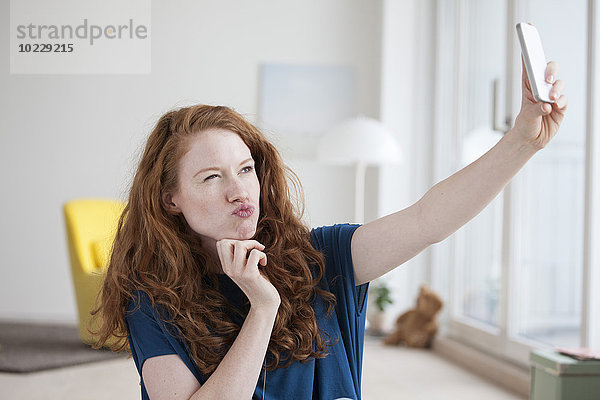 Junge Frau sitzt in ihrem Wohnzimmer und nimmt einen Selfie mit dem Smartphone.