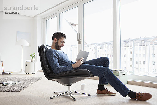 Junger Mann sitzt auf einem Ledersessel in seinem Wohnzimmer und benutzt ein digitales Tablett.