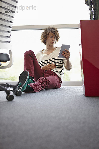 Junger Mann im Büro sitzend auf dem Boden mit digitalem Tablett