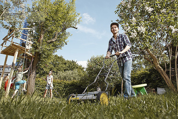 Mann mäht Rasen im Garten mit Familie im Hintergrund
