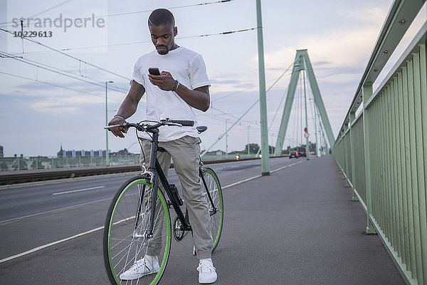 Deutschland  Köln  junger Mann mit Fahrrad auf der Rheinbrücke beim Blick auf sein Smartphone