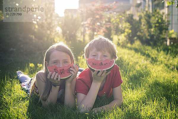 Junge und Mädchen liegen auf einer Wiese und bedecken einen Teil ihres Gesichts mit einer Scheibe Wassermelone.