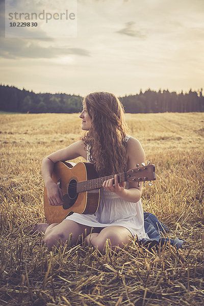 Junge Frau mit Gitarre am Abend auf Gerstenfeld sitzend
