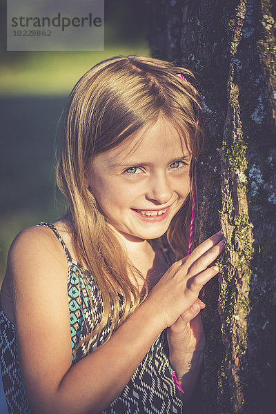 Porträt eines lächelnden Mädchens  das sich in der Abenddämmerung auf einen Baumstamm stützt.