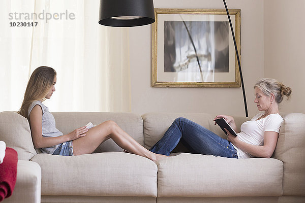 Mutter und Tochter sitzend mit digitalem Tablett und Buch auf der Couch