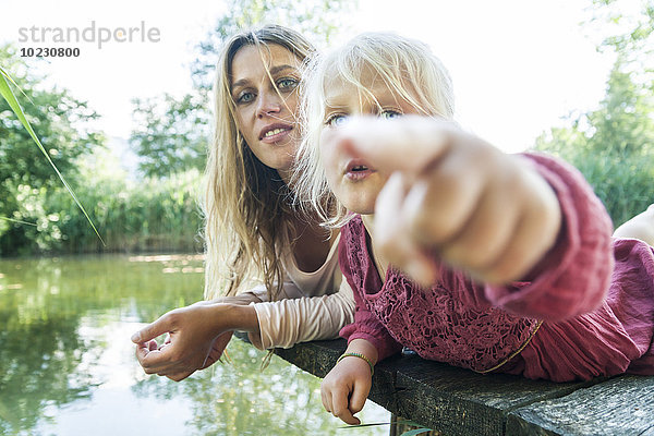 Mädchen liegt mit Mutter am Steg an einem See und zeigt mit dem Finger.