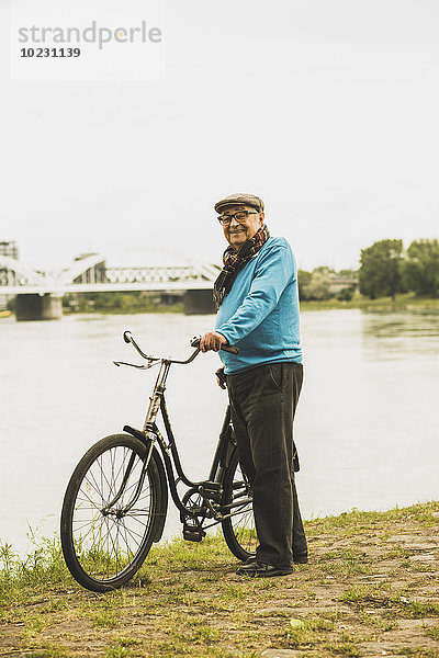 Porträt eines lächelnden älteren Mannes  der mit dem Fahrrad am Wasser steht.