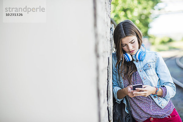 Brünette junge Frau mit Kopfhörer und Handy gegen die Wand gelehnt