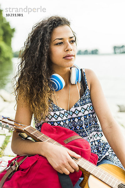 Junge Frau mit Kopfhörer  Gitarre und Rucksack am Flussufer