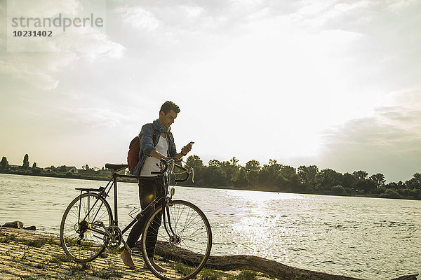 Junger Mann mit Fahrrad und Handy am Flussufer