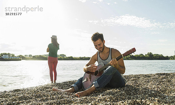 Junger Mann spielt Gitarre mit Frau am Flussufer