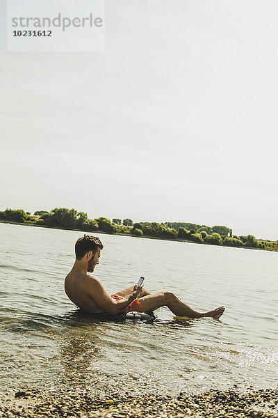 Junger Mann sitzt in einer Röhre im Fluss und benutzt ein Smartphone.