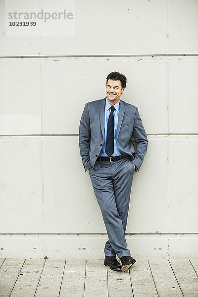 Porträt eines lächelnden Geschäftsmannes im grauen Anzug  der sich an die Betonwand lehnt.