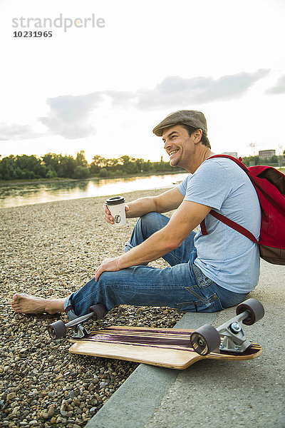 Lächelnder Mann sitzt am steinigen Strand mit Skateboard und Kaffee zum Mitnehmen.