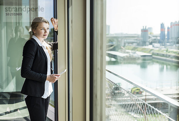 Junge Geschäftsfrau mit digitalem Tablett aus dem Fenster schauend
