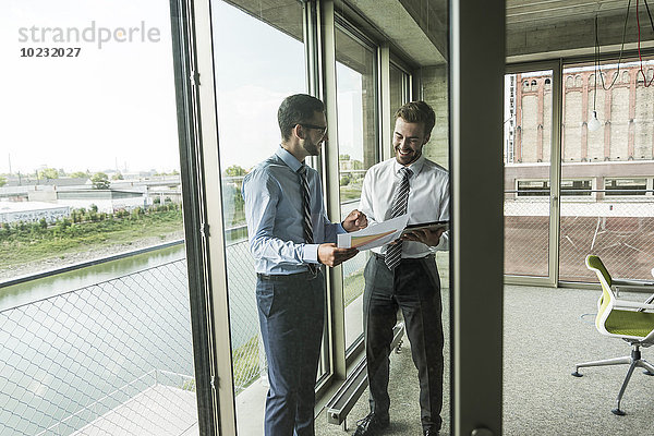Zwei junge Geschäftsleute schauen sich Dokumente und digitales Tablett am Fenster an.