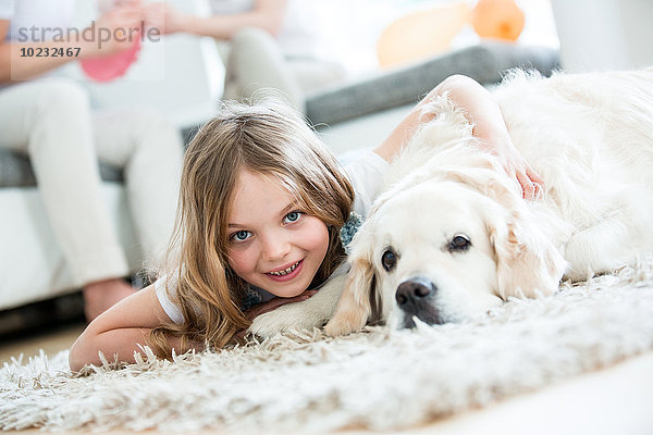 Kleines Mädchen kuschelt mit ihrem Hund  auf dem Boden liegend  Eltern im Hintergrund