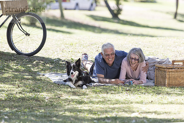 Lachendes Seniorenpaar mit Hund auf Decke auf einer Wiese liegend