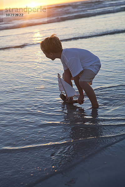 Junge spielt mit einem Spielzeug-Holzboot im Wasser am Strand