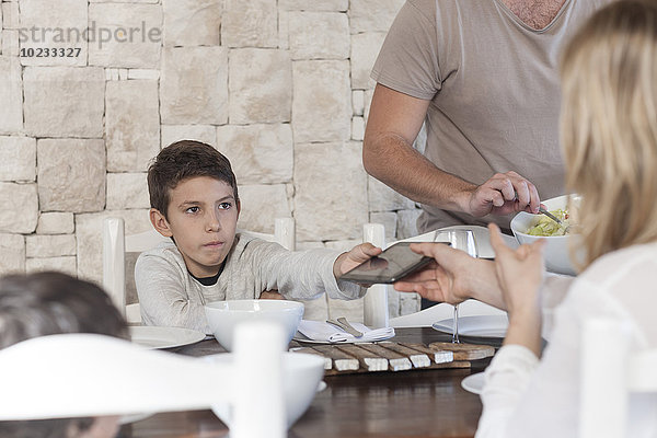 Familie beim Mittagessen am Esstisch mit dem Jungen  der die digitale Tafel der Mutter übergibt.