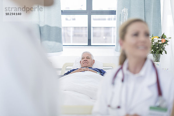 Älterer Mann schläft in einem Krankenhausbett  während die Ärzte im Vordergrund diskutieren.