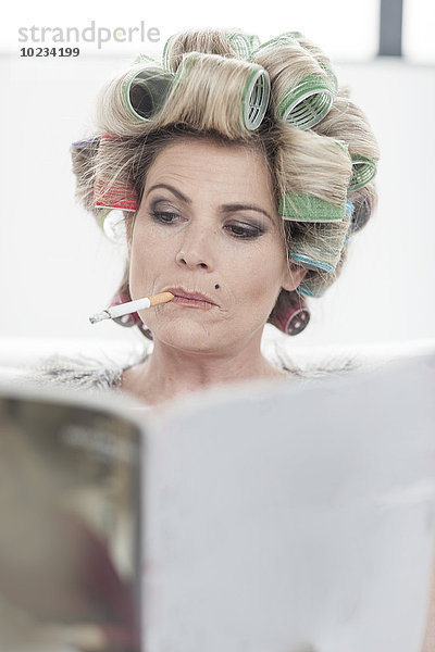 Porträt einer Frau mit Lockenwickler und Zeitschrift  die eine Zigarette raucht.