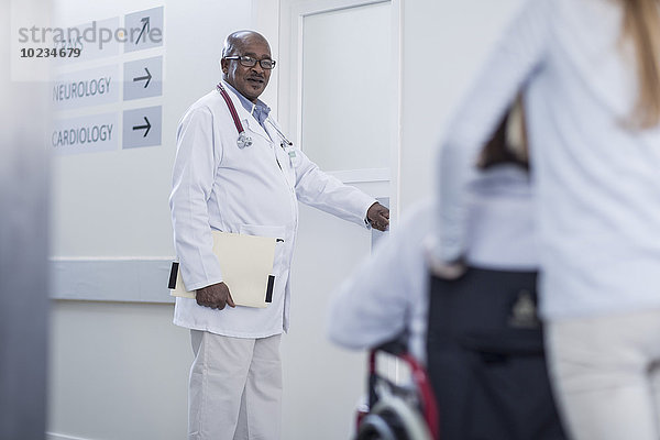 Arzt im Krankenhausflur öffnet Tür und Patient im Rollstuhl