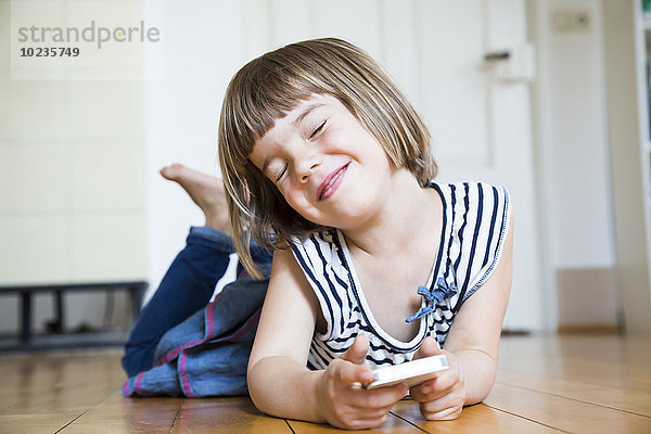 Lächelndes kleines Mädchen auf Holzboden liegend mit Smartphone