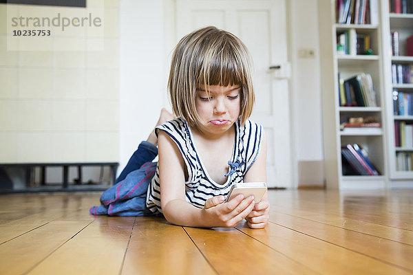 Kleines Mädchen liegt auf Holzboden und sieht skeptisch auf das Smartphone.