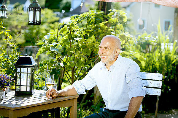 Porträt eines glücklichen Mannes  der mit einem Glas Weißwein im Garten sitzt.