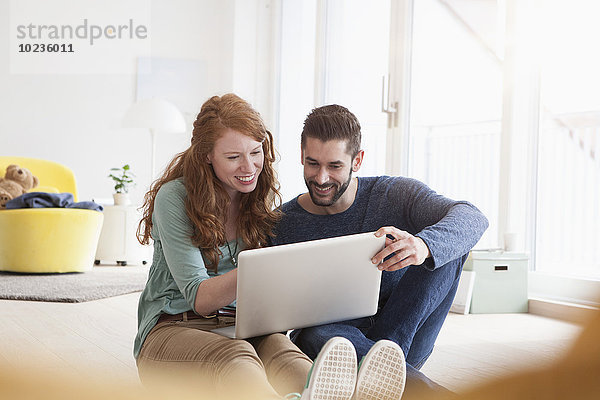 Lächelndes junges Paar sitzt auf dem Boden im Wohnzimmer mit Laptop