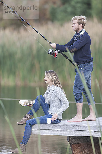 Junges Paar mit Buch  Tasse und Angelrute auf einem Steg am See