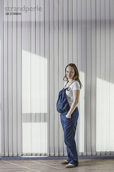 Ganzkörperporträt einer schwangeren Frau im Overall gegen den Vorhang stehend