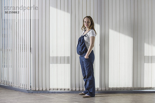 Ganzkörperporträt einer schwangeren Frau im Overall gegen den Vorhang stehend