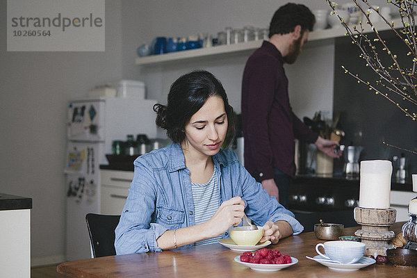 Junge Frau beim Kaffeetrinken am Küchentisch mit Mann im Hintergrund
