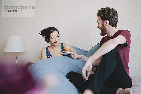 Porträt einer glücklichen jungen Frau  die einen Geburtstagskuchen mit Kerze hält  während sie neben einem Mann auf dem Bett sitzt.