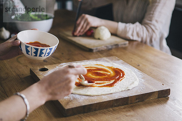 Abgeschnittenes Bild einer Frau  die Tomatensauce über Pizzateig streicht.